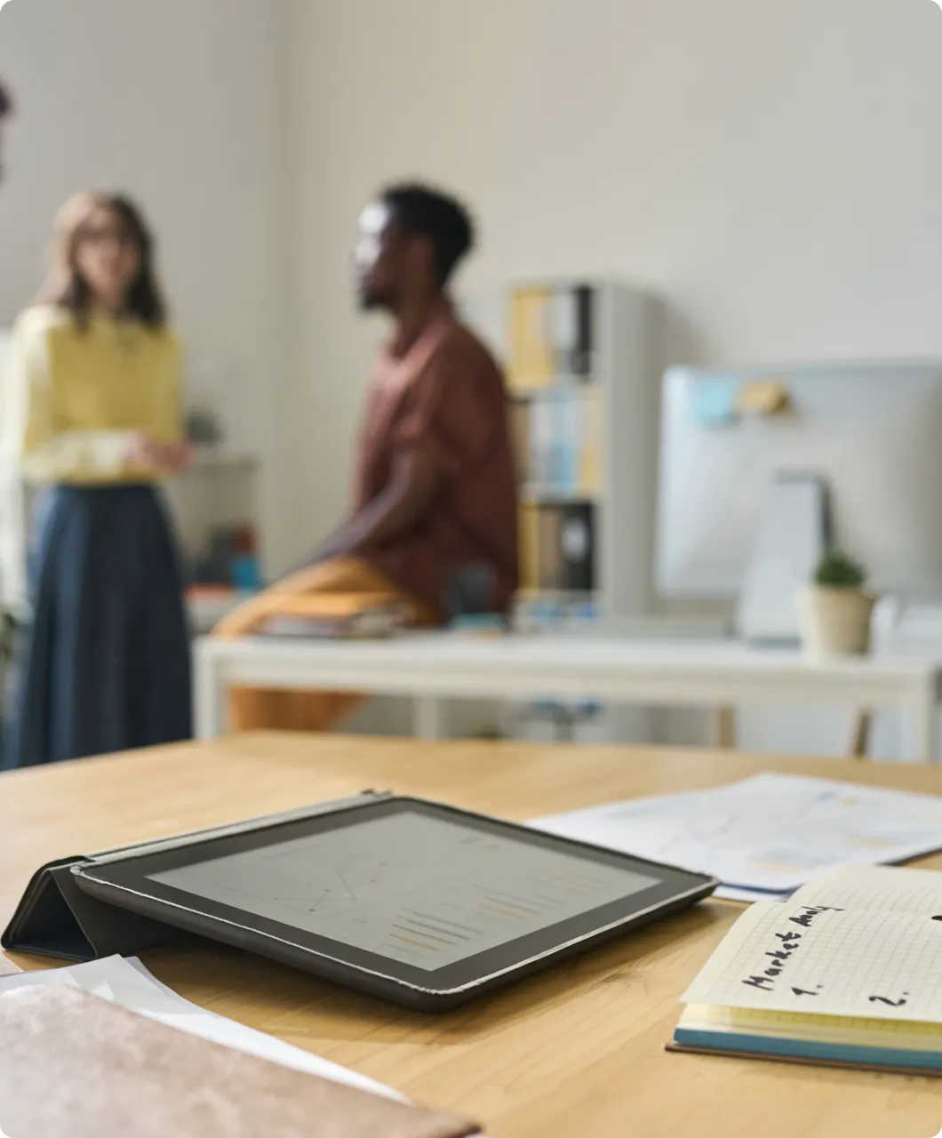 Tablet displaying charts on a desk with documents and a notebook labeled 'Market Analysis,' with two people conversing in the blurred background of an office.
