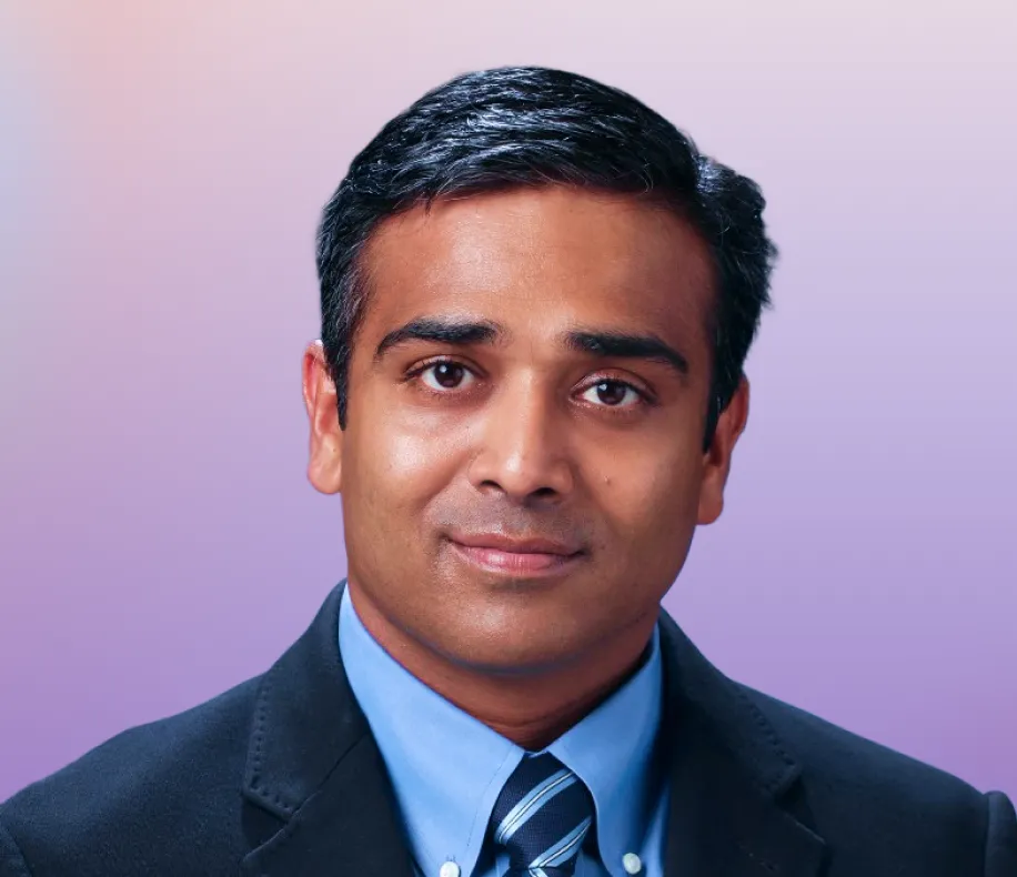 Portrait of a man wearing a dark blazer, blue shirt, and striped tie against a purple gradient background.