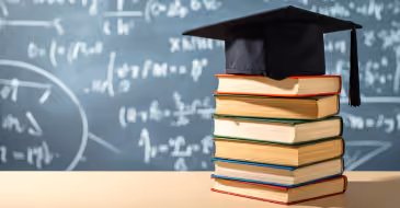 A stack of books with a graduation cap on top, sat on a desk in front of a blackboard