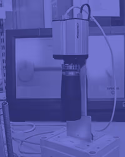 Close-up of a laboratory testing device with a cylindrical component and wires connected, positioned on a white platform with screens in the background.