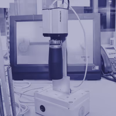 Close-up of a precision measurement device with a mounted lens and a computer monitor in the background.