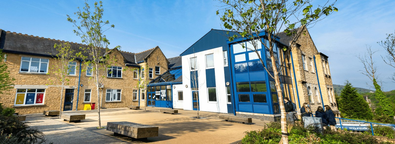 Sunny courtyard at Bacup and Rawtenstall Grammar School in Waterfoot, with stone school buildings, a modern blue and white extension, benches and young trees