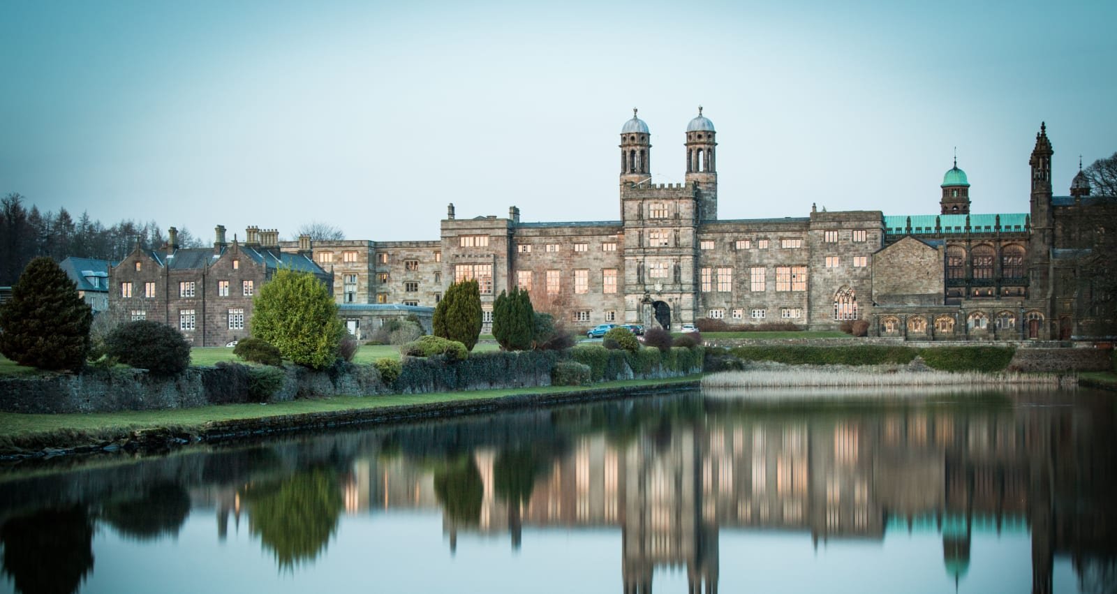 Stonyhurst College's long stone front with twin domed towers, reflected in the still water of a lake, surrounded by trees and hedges
