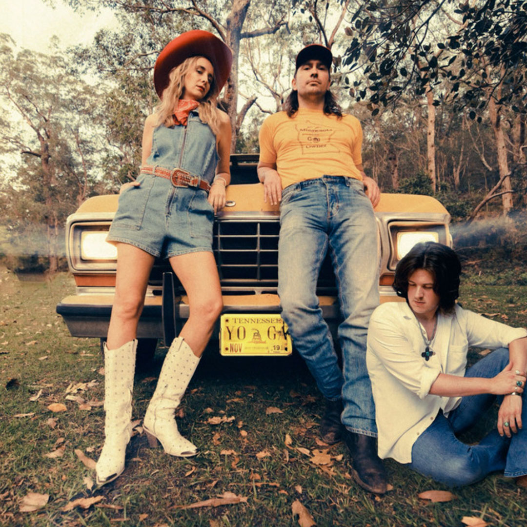 Three young adults posing outdoors in front of a vintage yellow truck with Tennessee license plate, surrounded by trees.