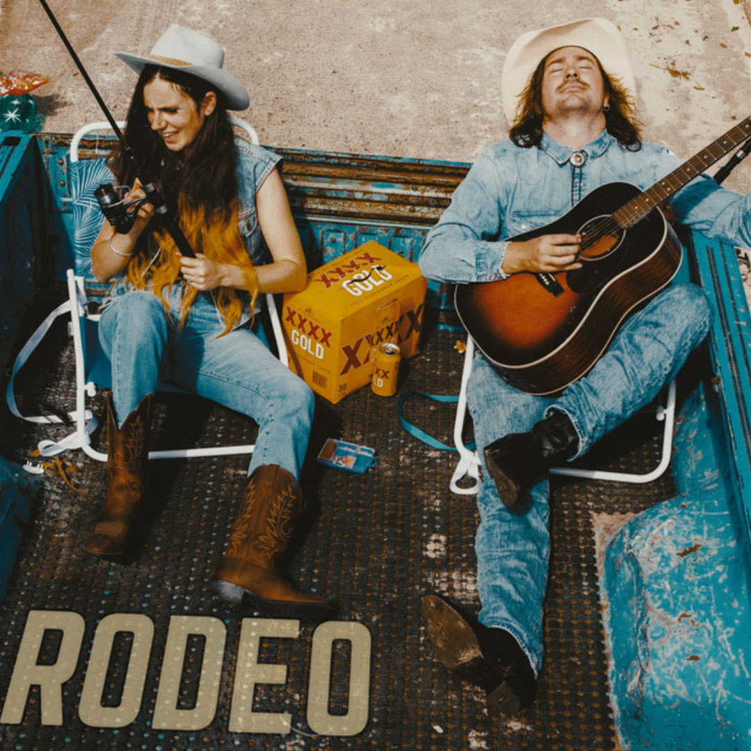 Two people in denim and cowboy hats sitting in the back of an old blue truck; one is playing guitar while the other holds a fishing rod, with a box and can of beer between them on the truck bed.