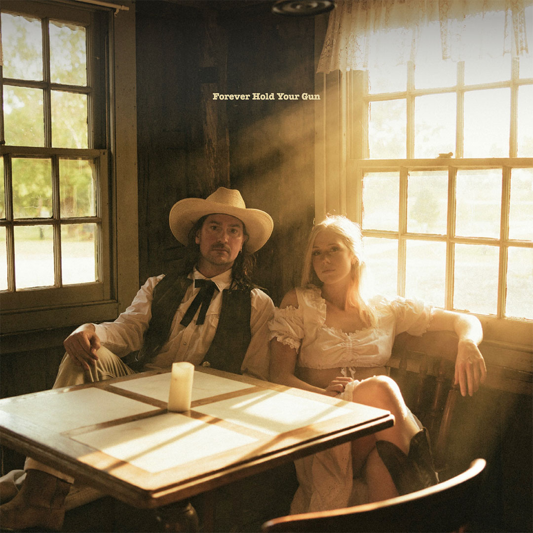 A man in a white cowboy hat and vest and a woman in a white vintage dress sit together at a wooden table inside a sunlit rustic room with large windows.