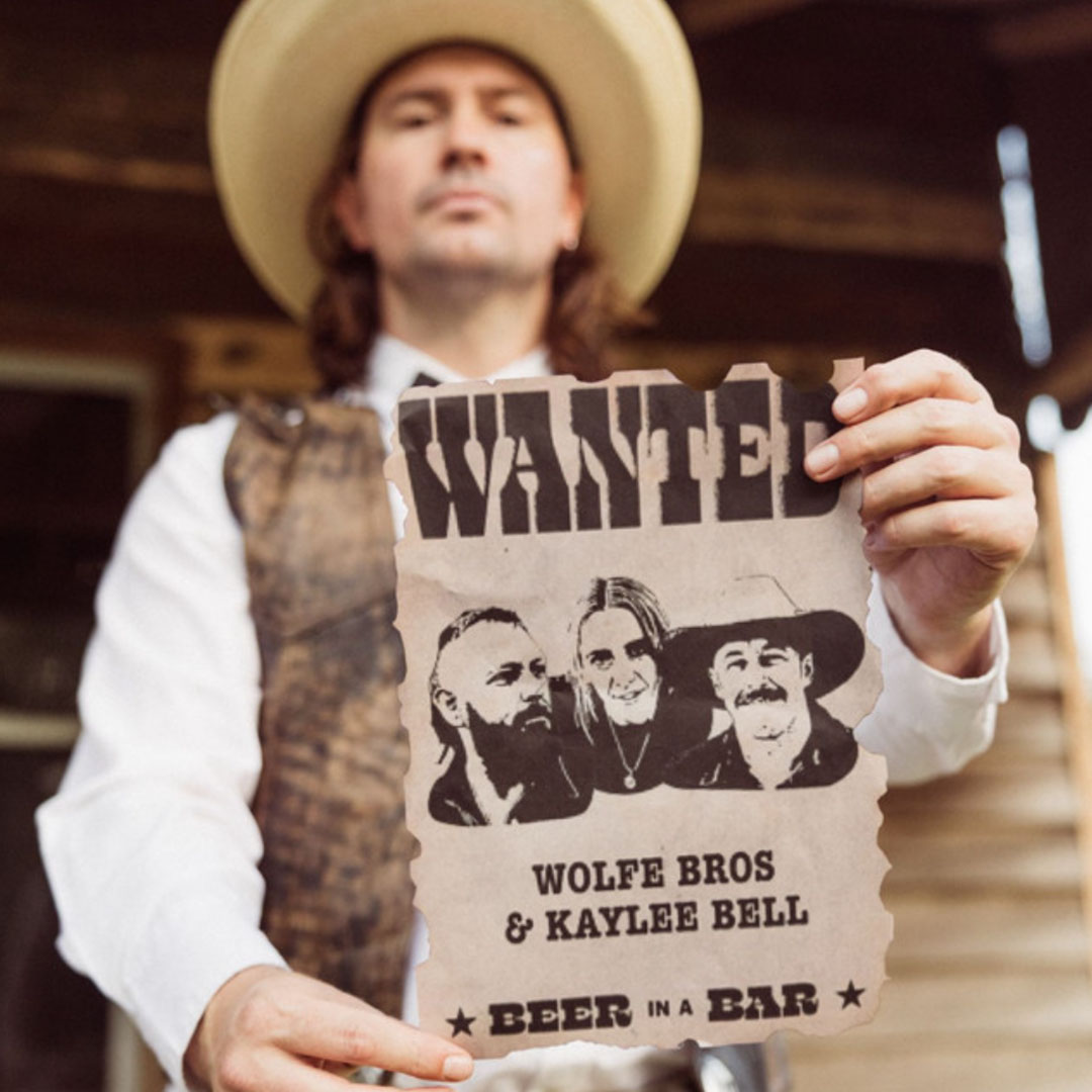 Man in cowboy hat holding a wanted poster featuring portraits of three people labeled Wolfe Bros & Kaylee Bell with text 'Beer in a Bar'.