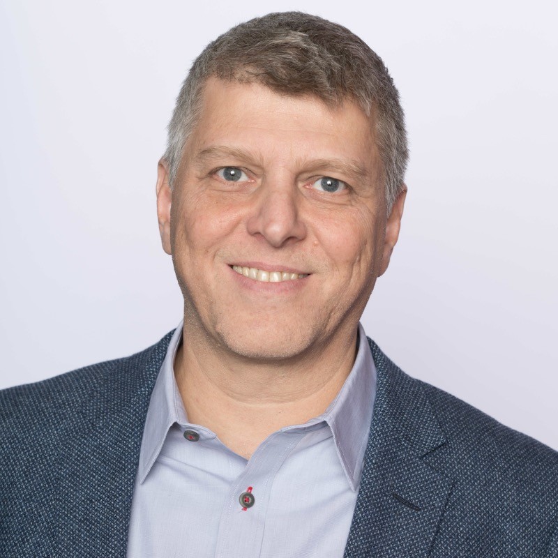 Smiling middle-aged man with short gray hair wearing a blue blazer and light blue shirt.