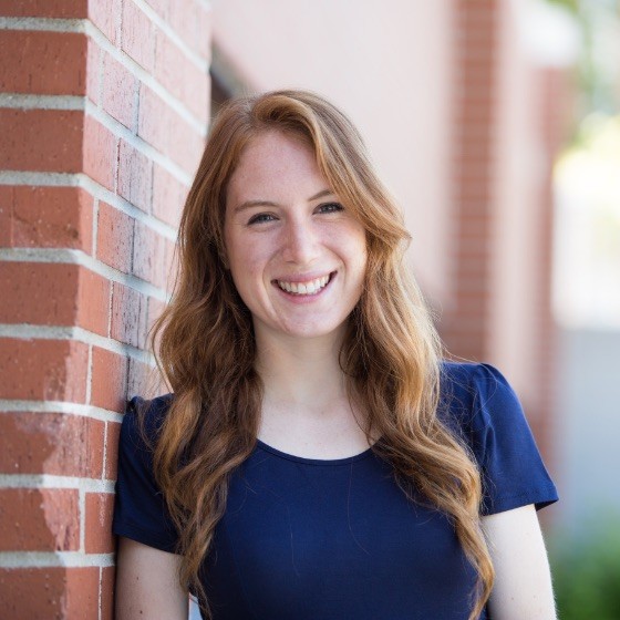 Smiling young woman with long red hair wearing a navy blue shirt leaning against a brick wall outdoors.