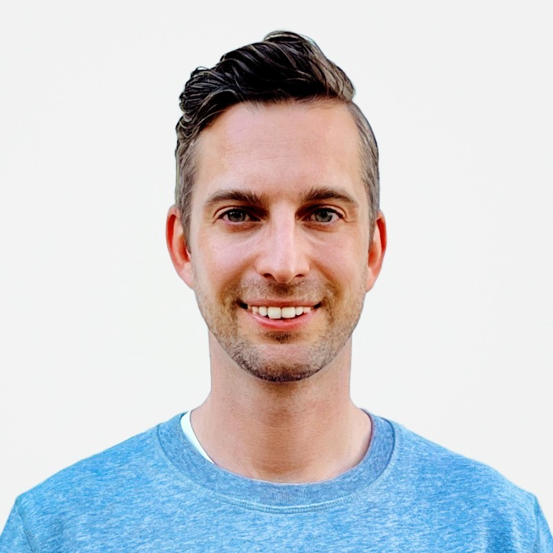 Smiling man with short dark hair wearing a blue crew-neck shirt against a plain white background.