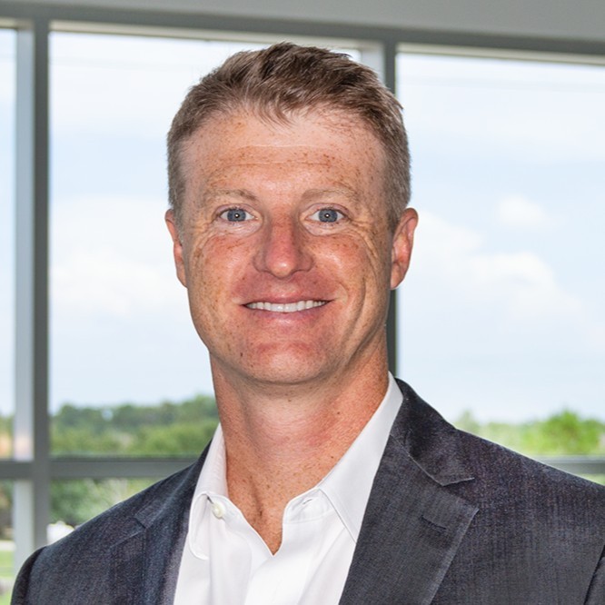 Smiling man with short light brown hair, wearing a dark suit jacket and white shirt, standing indoors with a window and greenery in the background.