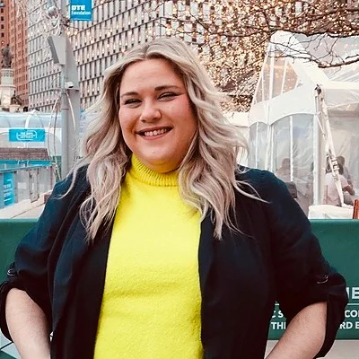 Smiling woman with wavy blonde hair wearing a bright yellow top and dark jacket standing outdoors in an urban setting.