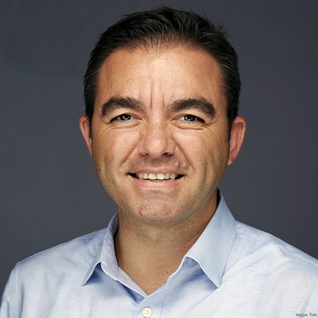 Smiling man with short dark hair wearing a light blue button-up shirt against a gray background.