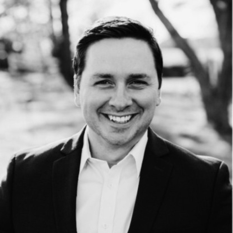 Smiling man in a suit jacket and white shirt outdoors with blurred trees in background, black and white photo.