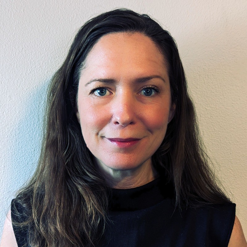 Woman with long brown hair, blue eyes, and a slight smile wearing a black sleeveless top against a plain light-colored wall.