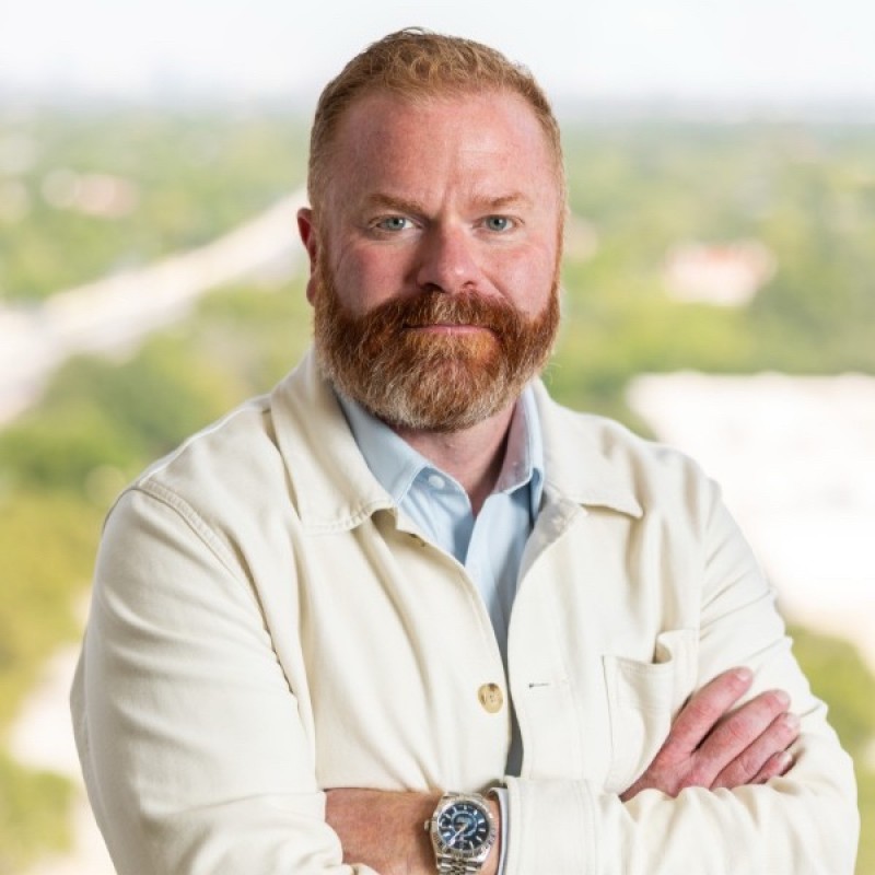 Bearded man with red hair wearing a white jacket and light blue shirt, standing with arms crossed against a blurred outdoor background.