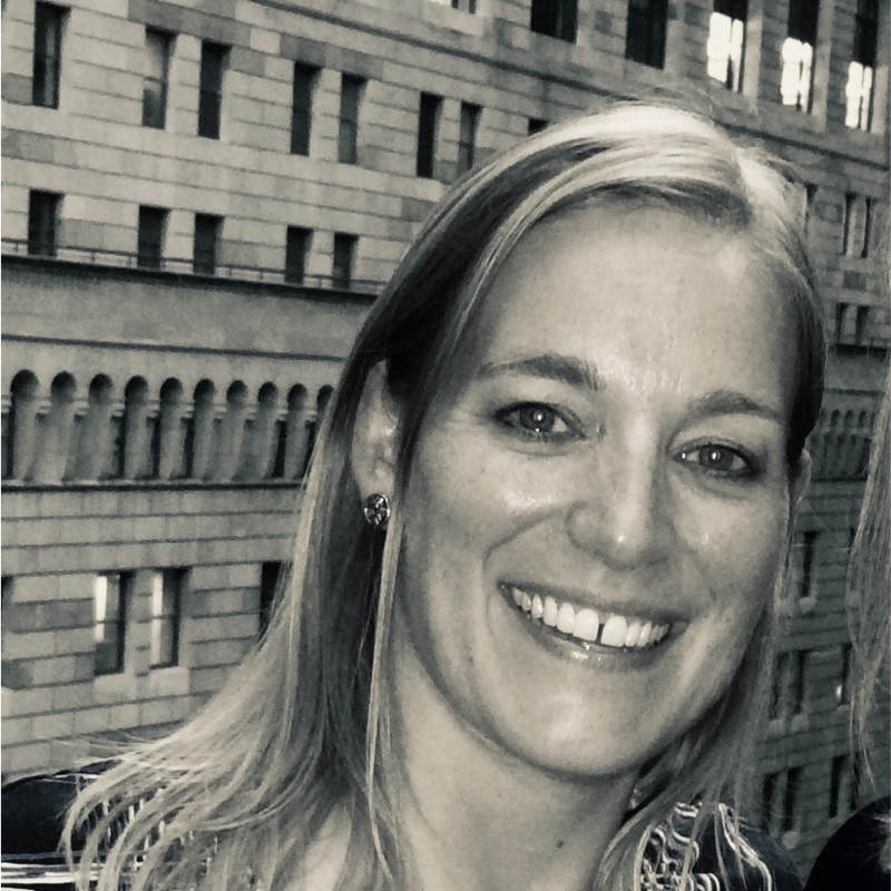 Black-and-white close-up of a smiling woman with light hair and earrings in front of a building facade.