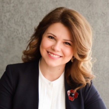 Smiling woman with shoulder-length light brown hair wearing a black blazer and white blouse against a gray background.