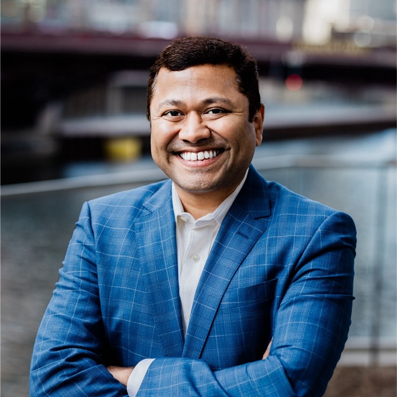 Smiling man in a blue checked suit with arms crossed standing outdoors near a waterfront.