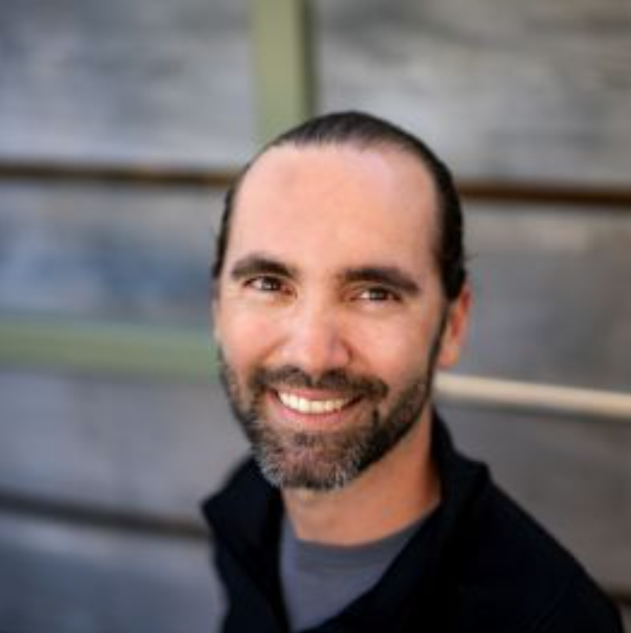 Smiling man with dark hair pulled back and a beard wearing a black jacket.