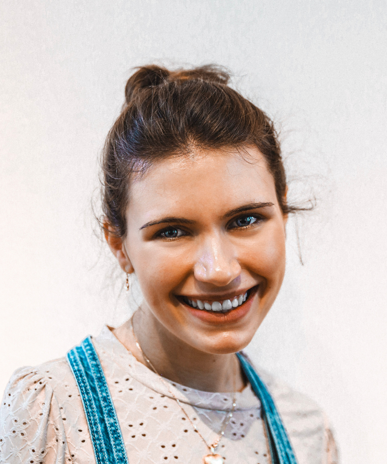 Smiling young woman with brown hair tied up, wearing a white eyelet blouse and denim overalls.