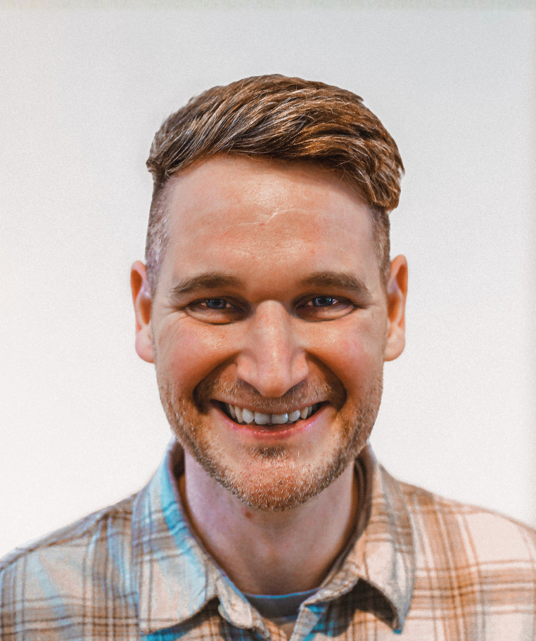 Smiling man with short brown hair and a trimmed beard wearing a plaid shirt against a plain background.