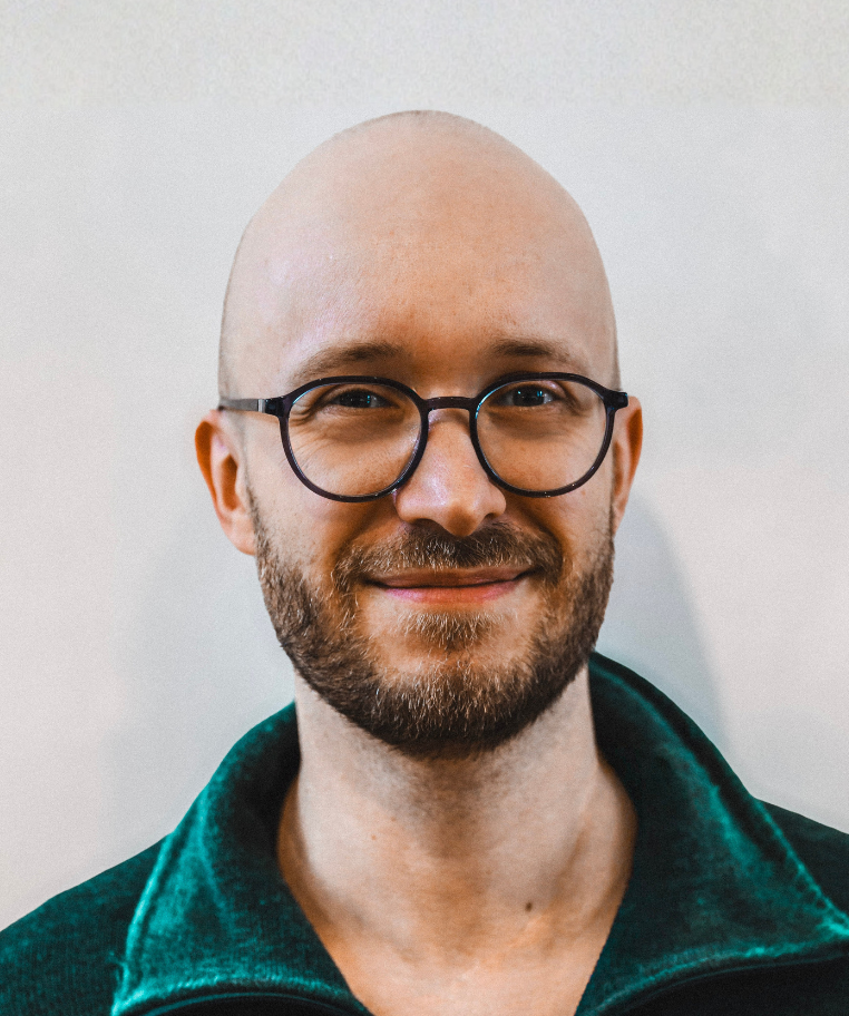 Smiling bald man with a beard and round glasses wearing a green jacket.