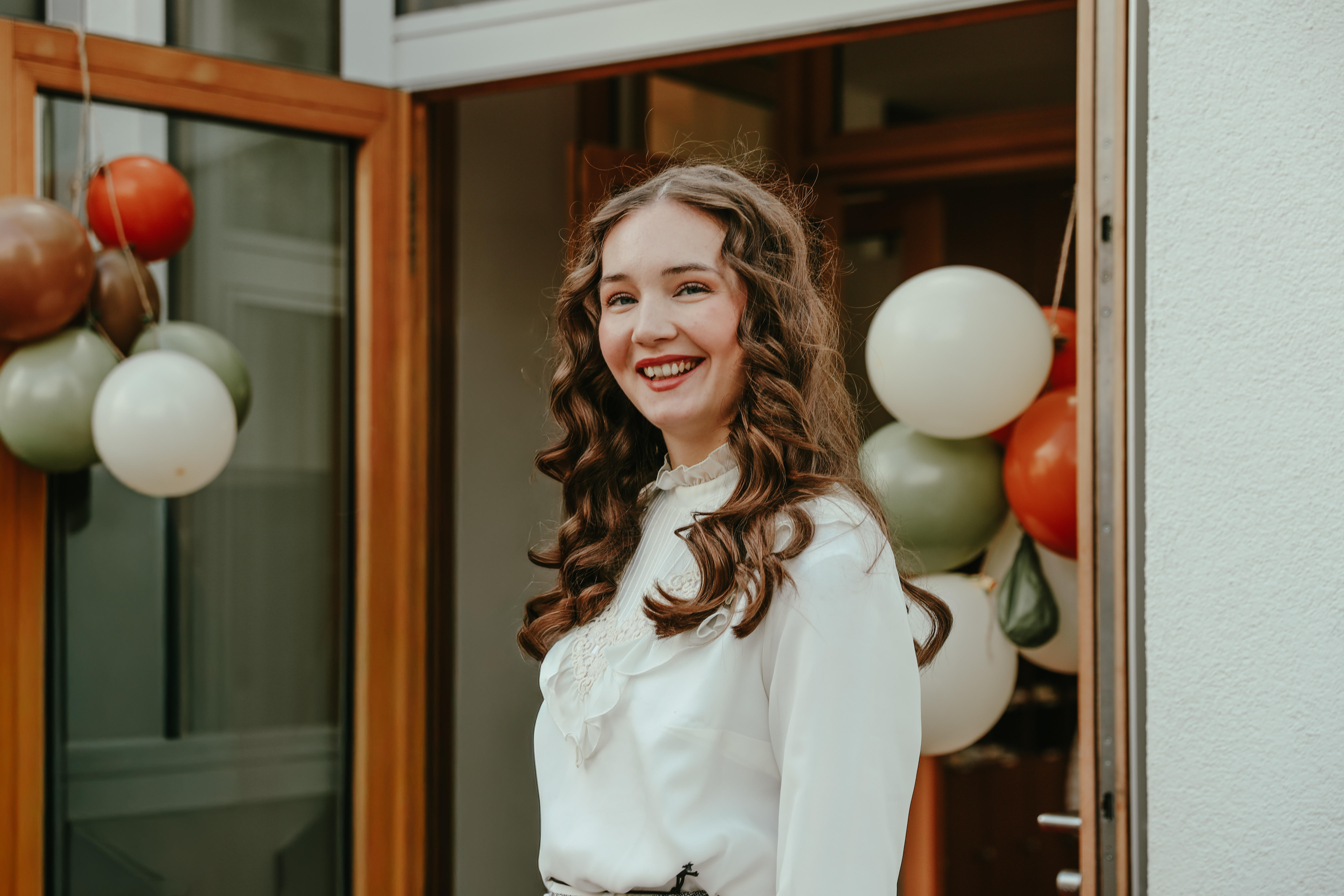 Young woman with long curly hair in a white blouse smiling near balloons by a wooden door.