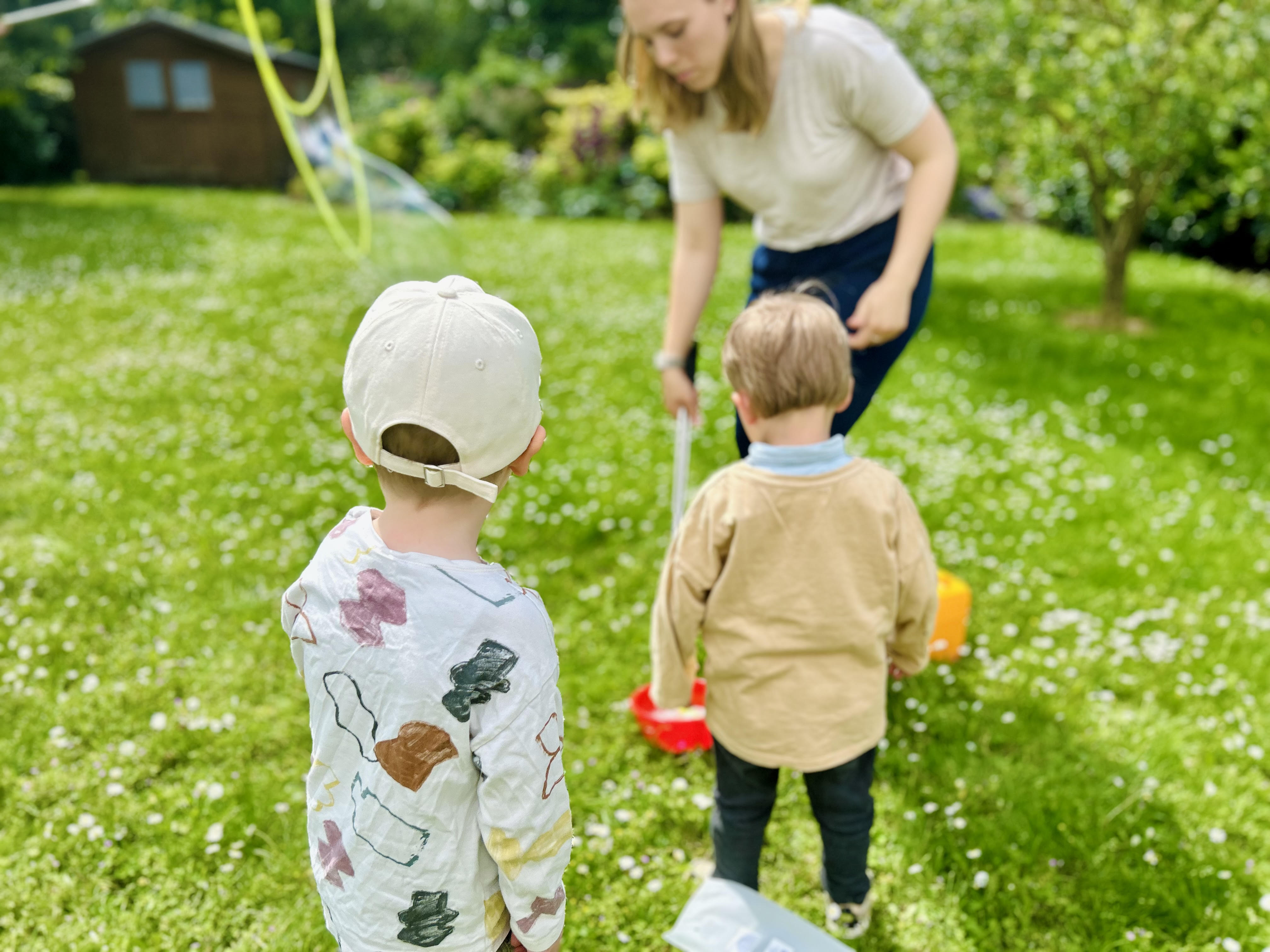 Two boys standing on grass watching a woman crouch down with a toy or tool in a green backyard.