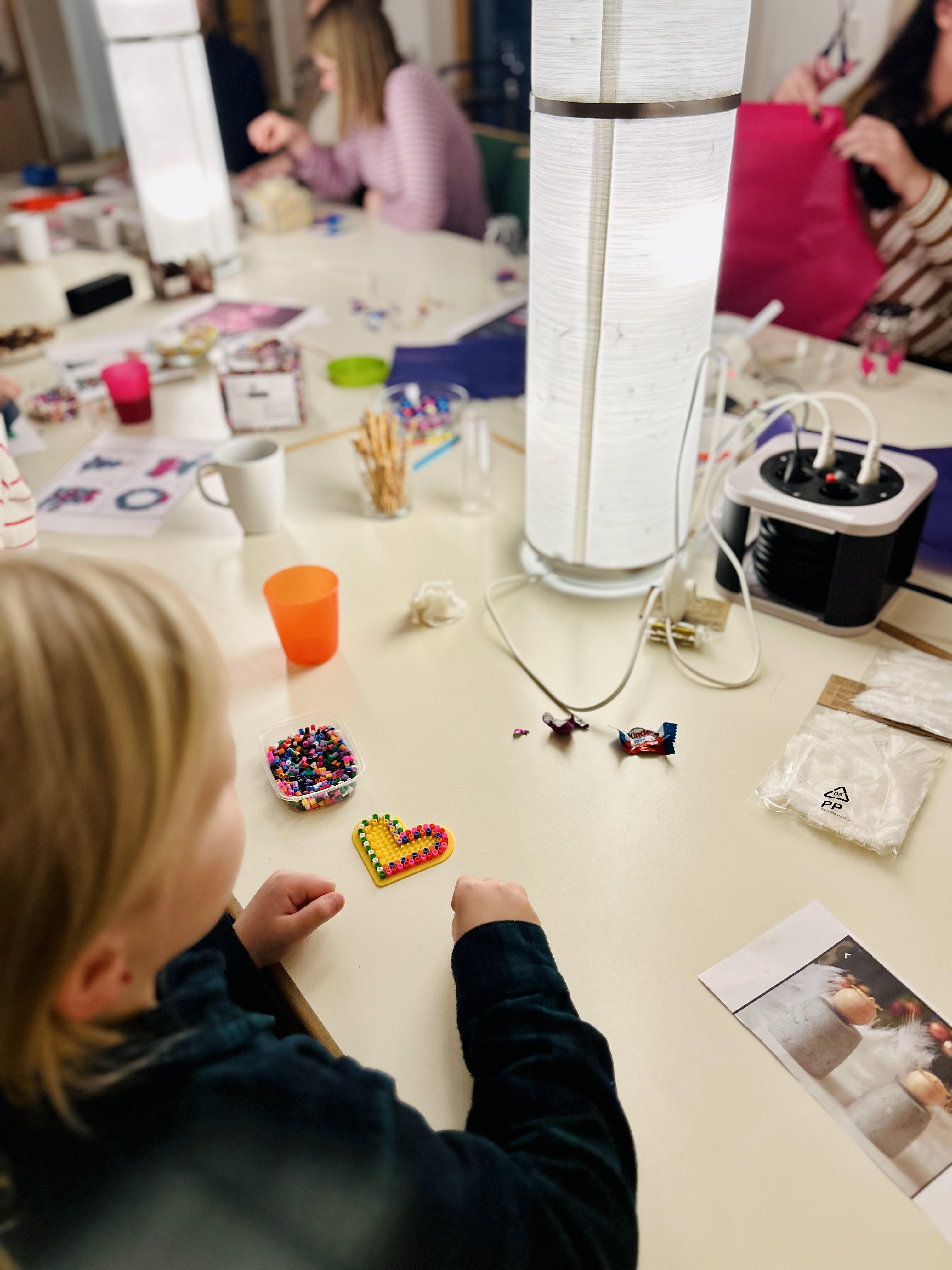 Child seated at a table crafting a heart shape with colorful beads and surrounded by crafting supplies.