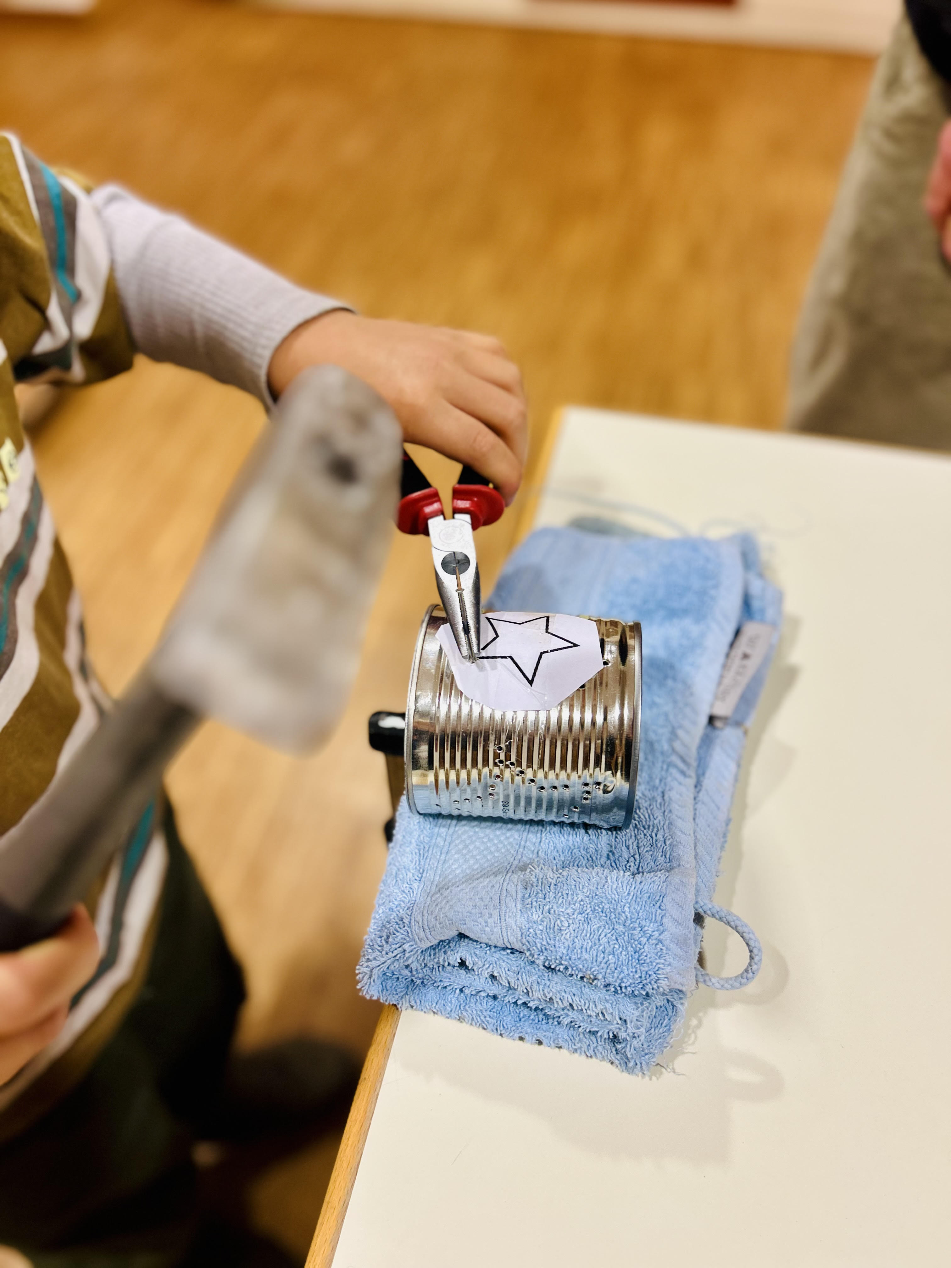 Child using pliers to punch holes in a tin can with a star paper stencil on a blue towel.