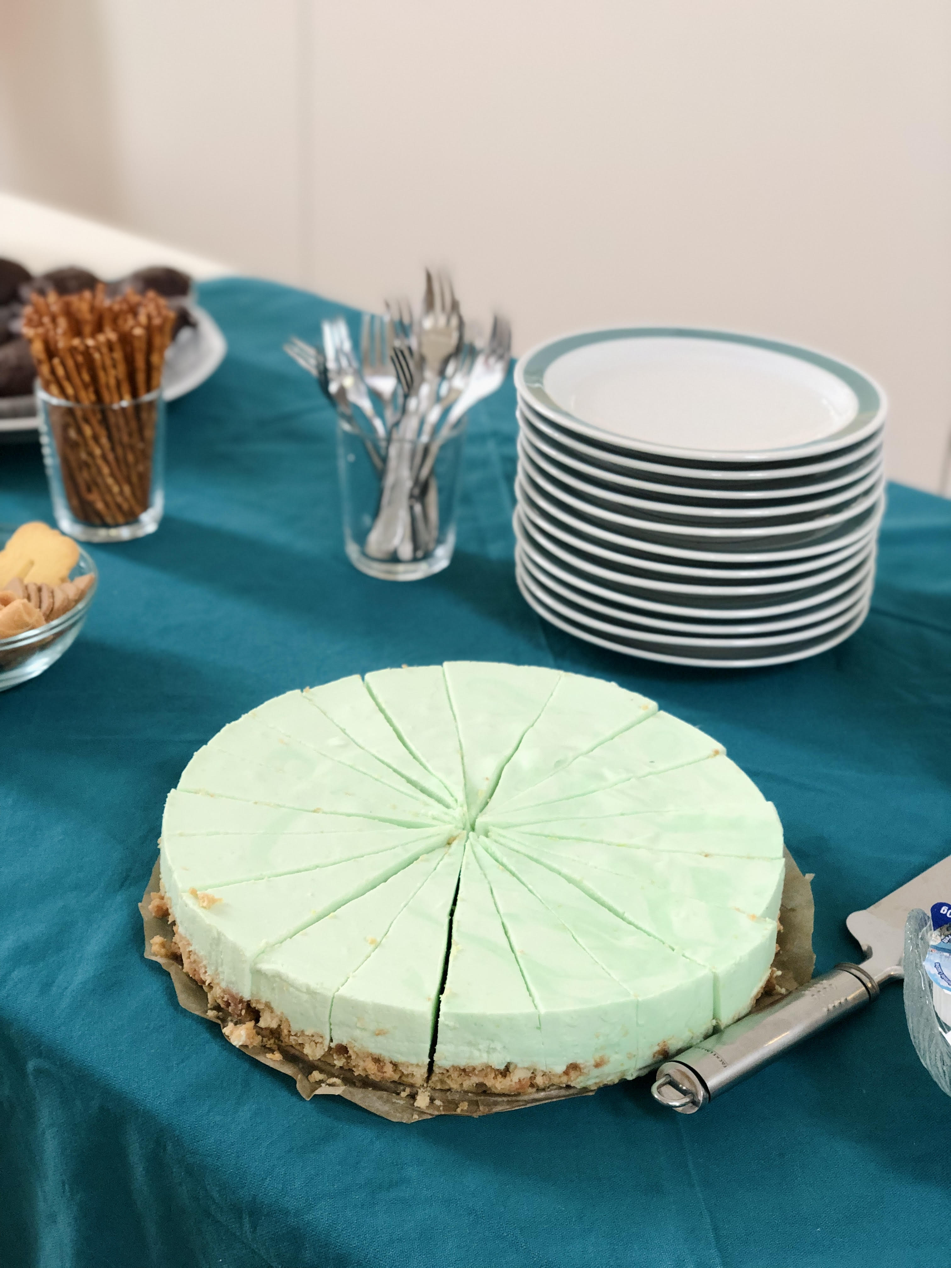 Round green cheesecake sliced into pieces on teal tablecloth with plates, forks, and snacks nearby.