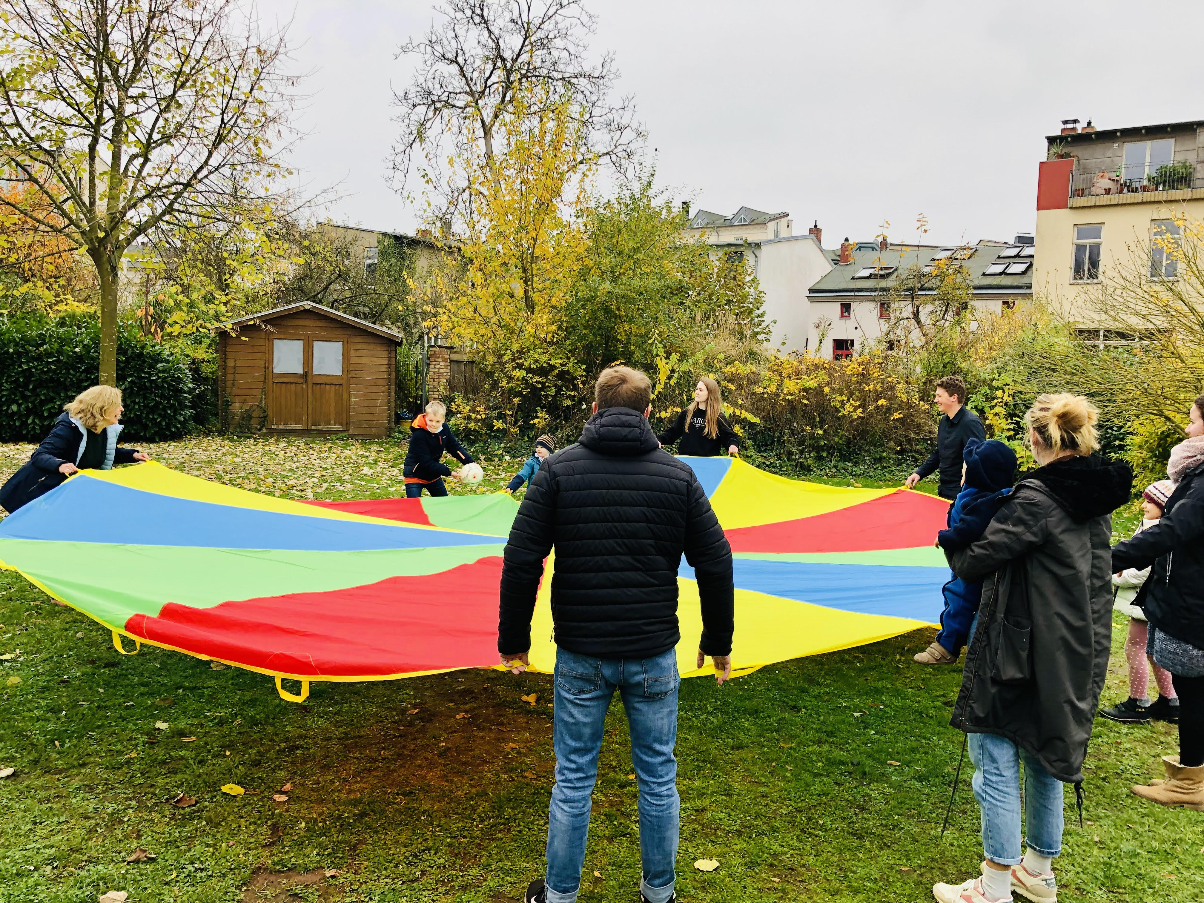 Group of adults and children outdoors holding colorful parachute in a backyard with autumn trees and shed.