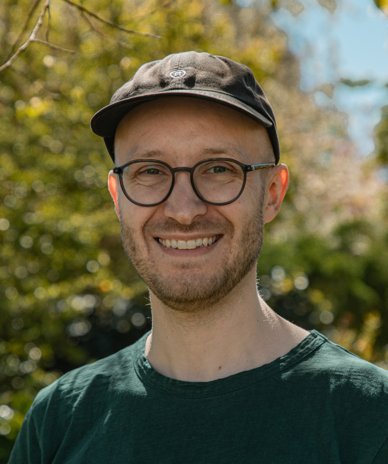 Smiling bald man with a beard and round glasses wearing a green jacket.