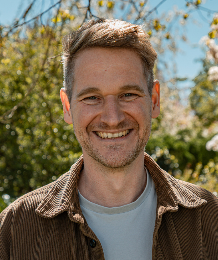 Smiling man with short brown hair and a trimmed beard wearing a plaid shirt against a plain background.