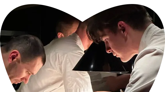 Three men in white shirts intently working or studying under a black angled desk lamp in a dimly lit setting.