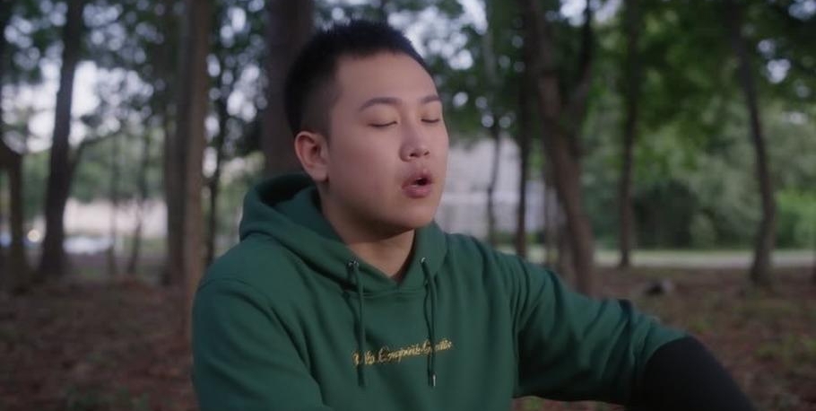 A Chinese American man with short hair and a green hoodie sits in a forest, exhaling after taking a deep breath.