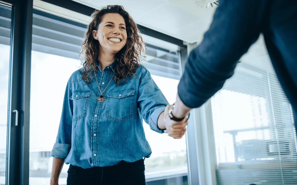 A woman smiling as she shakes hands with someone in a bright, modern office, suggesting the start or conclusion of a meeting or interview.