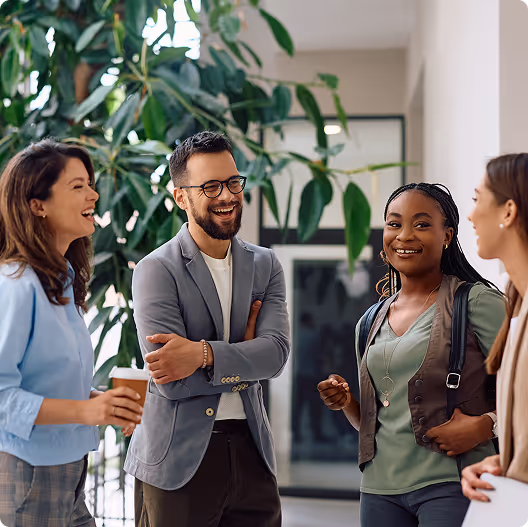 Four coworkers standing in an office space, smiling and chatting together, with one holding a coffee cup and plants in the background.
