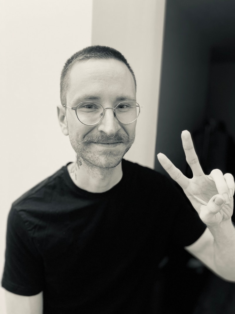 Hey, its me! I'm wearing a black t-shirt. In this photo, I have my hair shaved and am flashing a peace sign. I wear round, silver glasses and have a large flower tattoo on the right side of my neck.