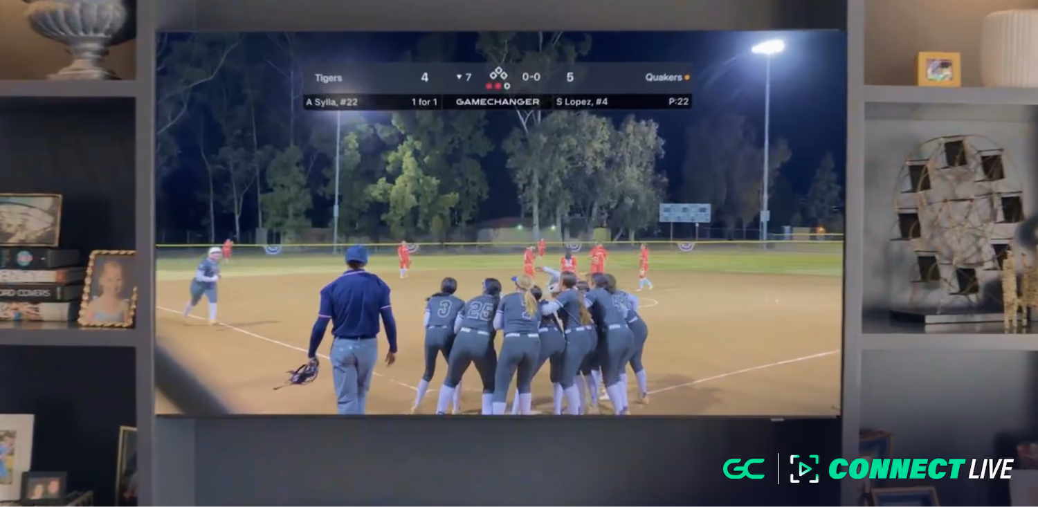 A group of softball players in gray uniforms and an umpire watch a play on a lit softball field at night.