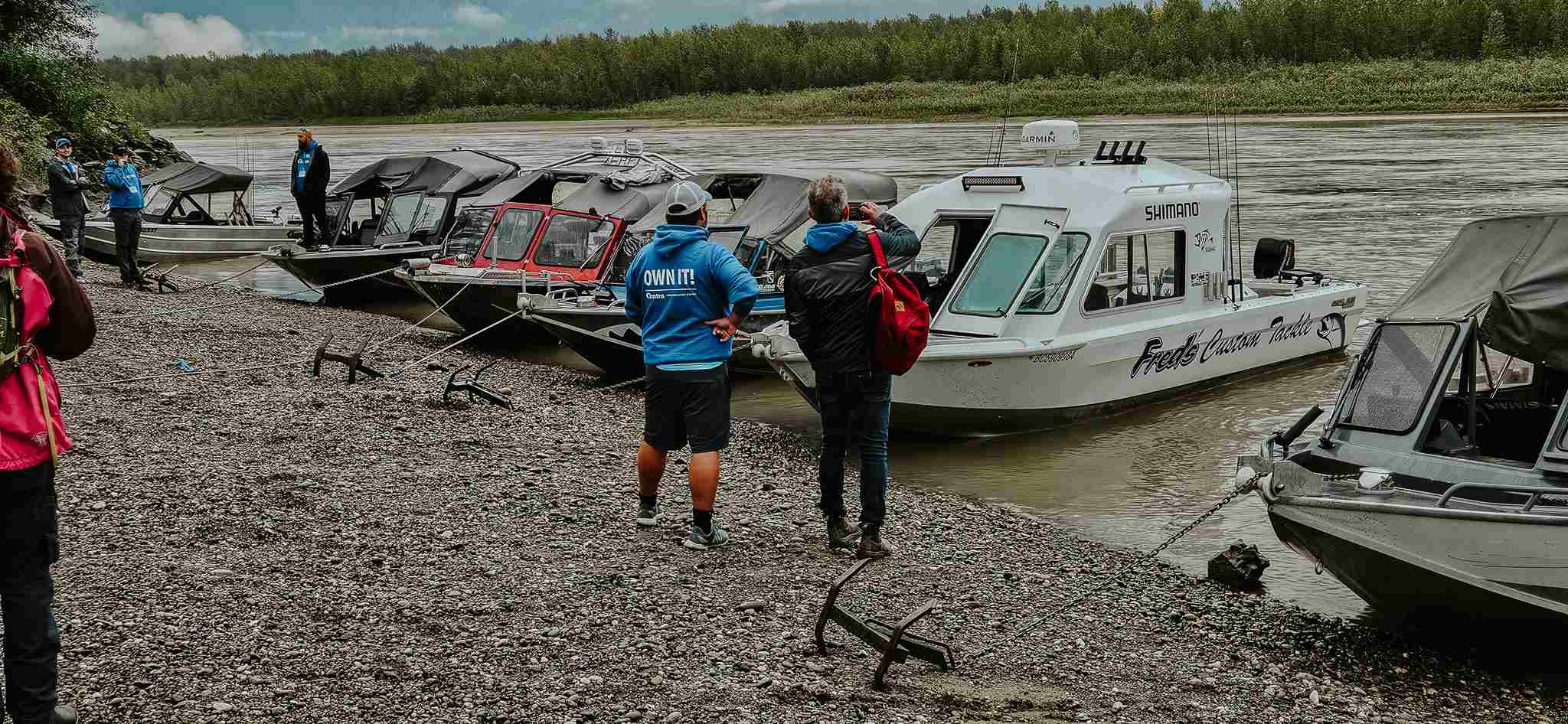 Guided fishing jet boats pulled up on a gravel shore of the Fraser River as guests in blue hoodies gather on the bank.