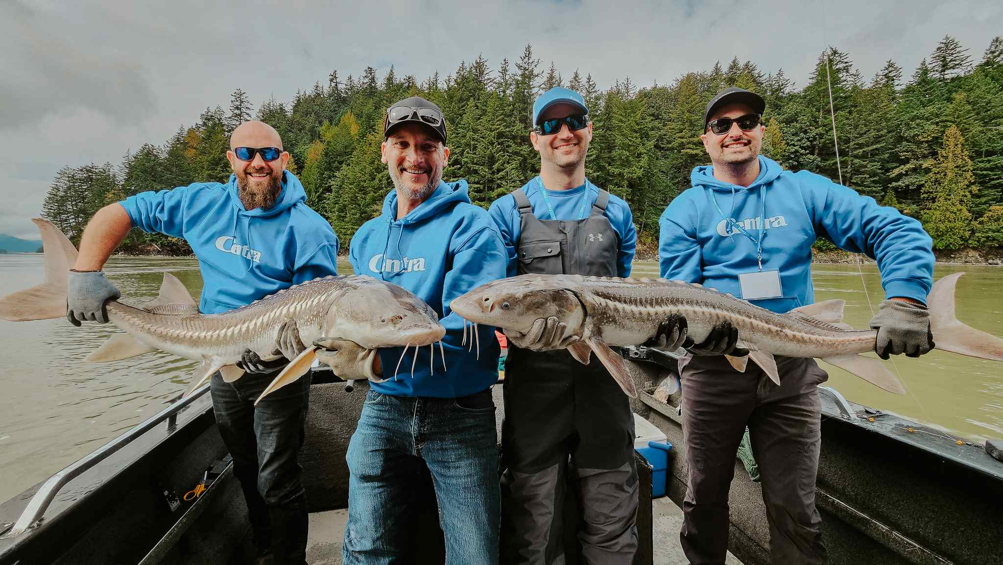 Four smiling anglers on a boat hold up two sturgeon between them, with evergreen forest behind.