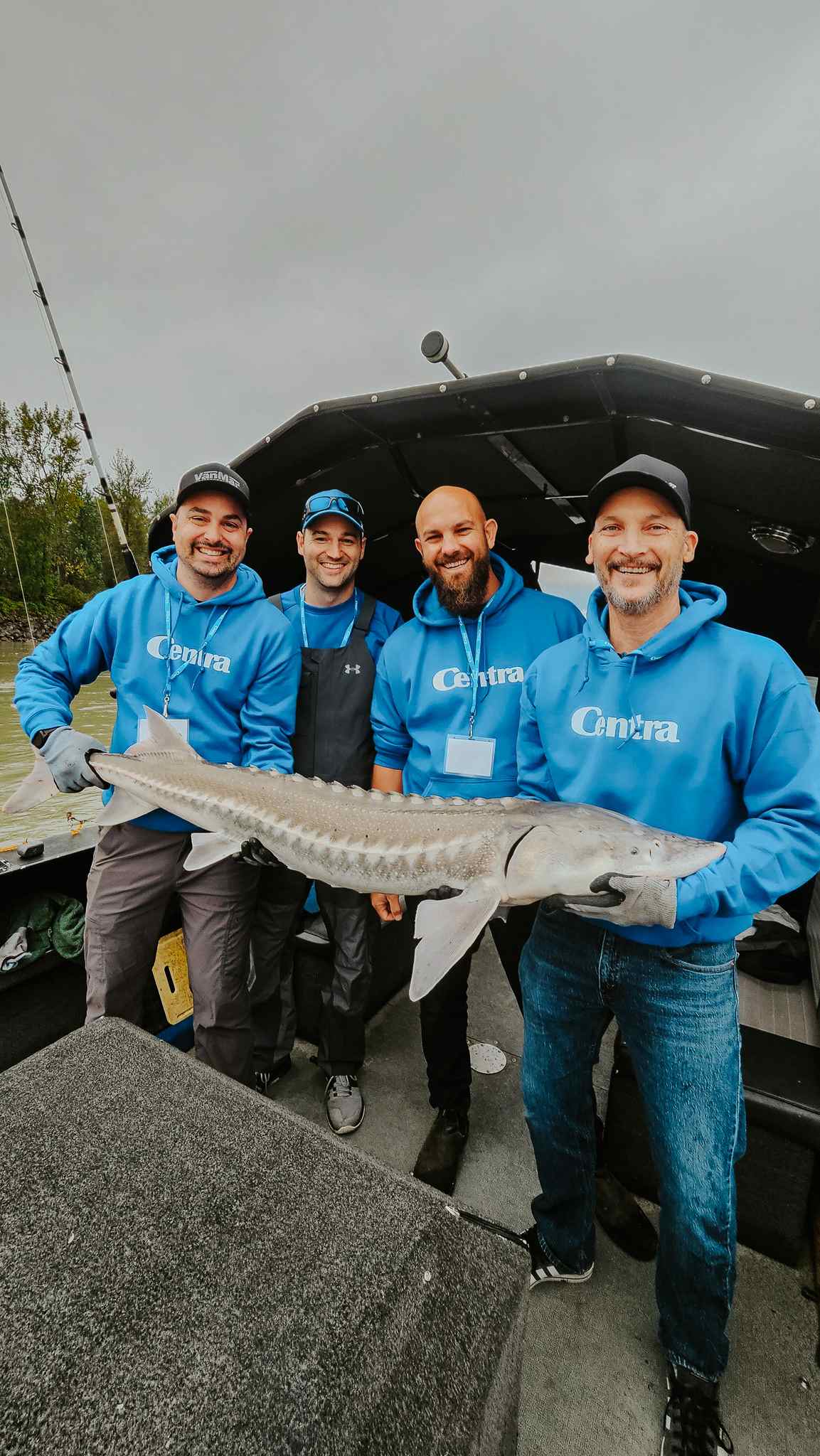 Four smiling anglers in matching blue hoodies pose on a fishing boat with a sturgeon.
