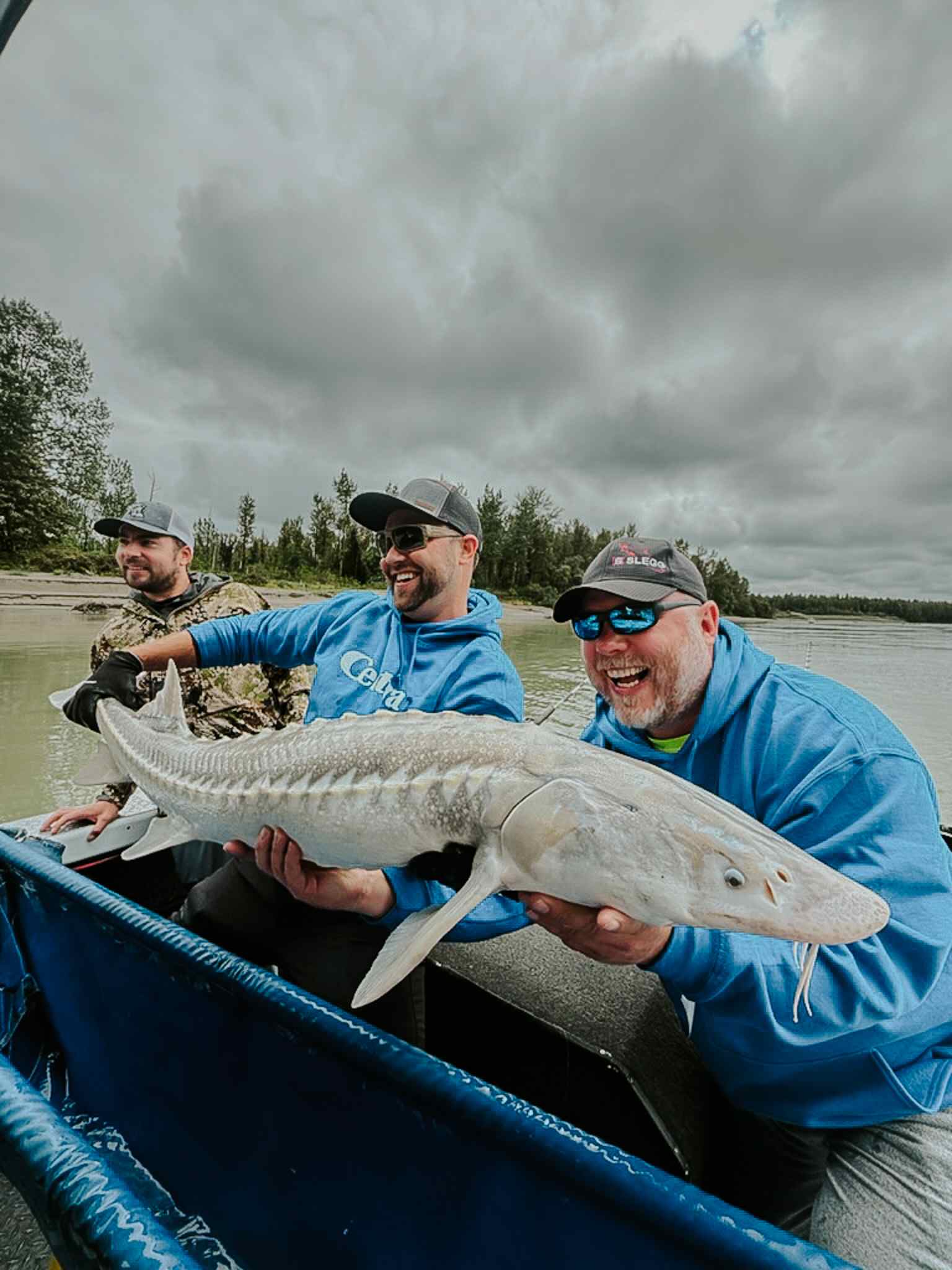 Three laughing anglers on a boat show off a pale sturgeon under a dramatic cloudy sky.