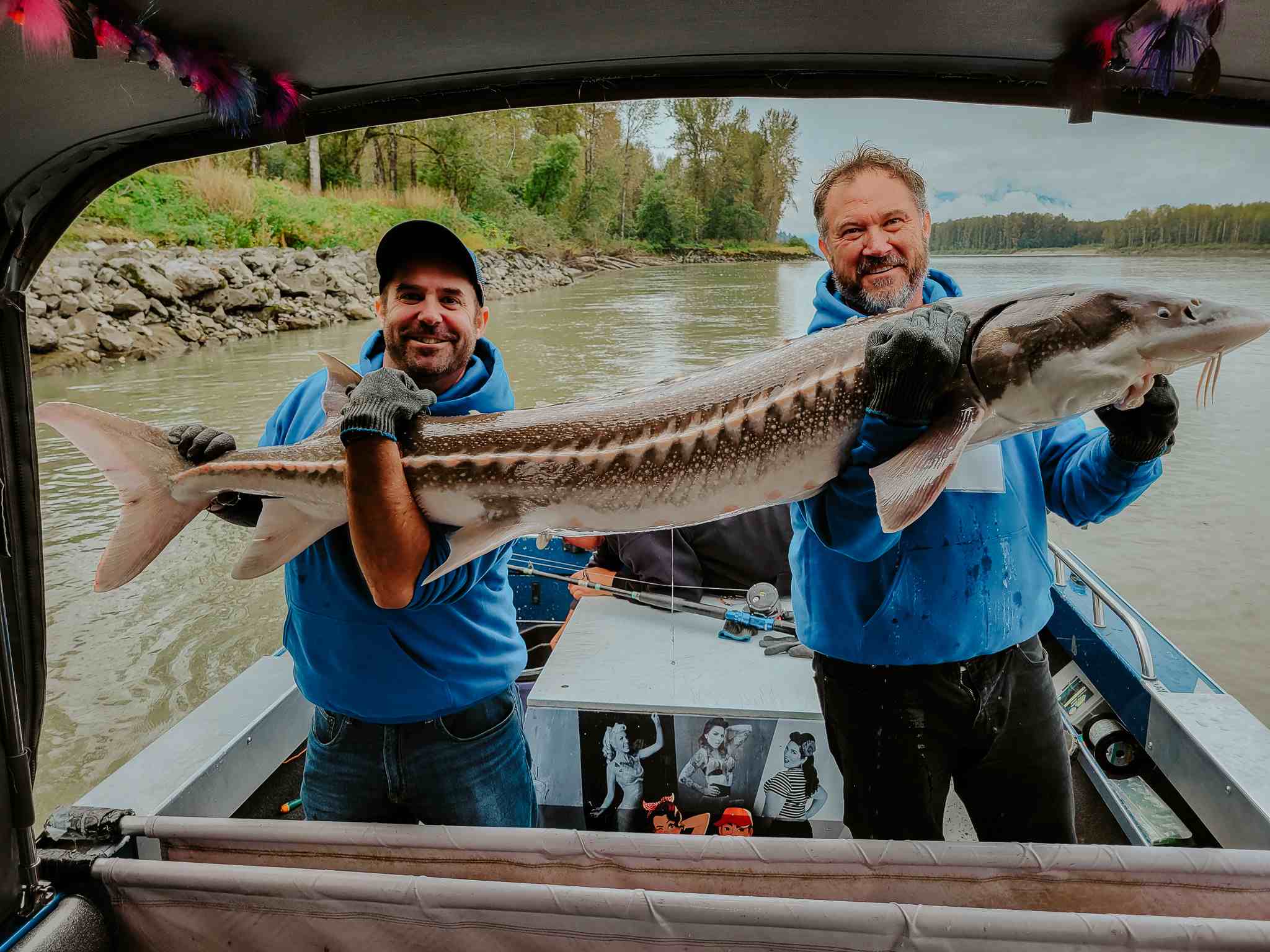 Two anglers hold a long sturgeon under a boat canopy, with the river and trees behind them.