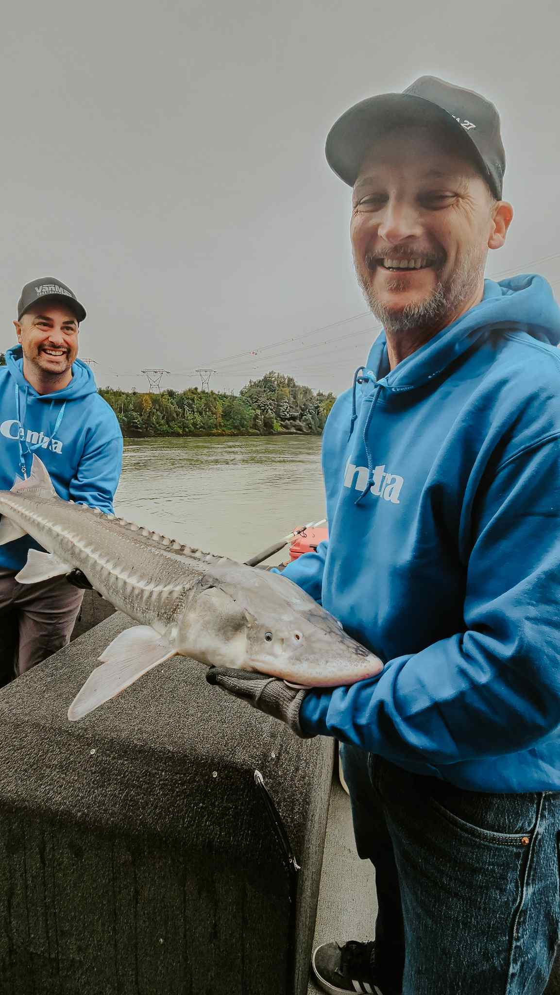 Two smiling anglers in blue hoodies hold a small sturgeon on a boat, with the Fraser River behind them.