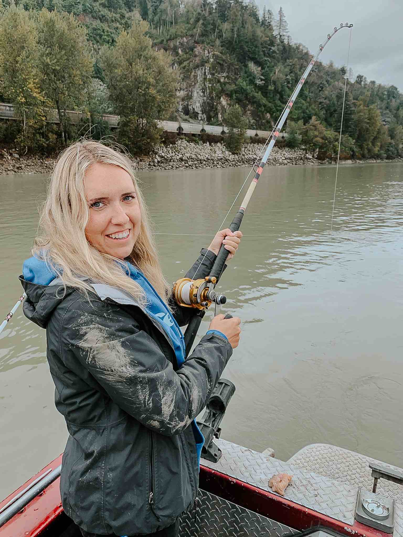 A smiling angler in a rain jacket holds a bent fishing rod on a boat, with a rocky bank and forested hillside behind her.