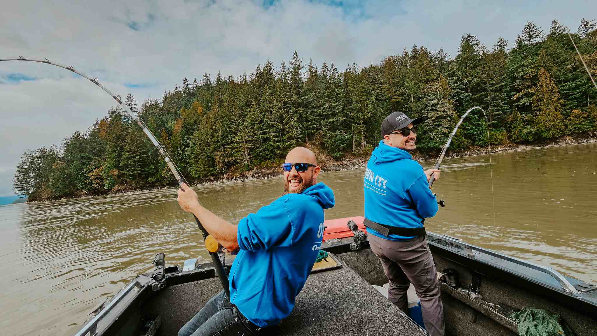 Two anglers laugh as their fishing rods bend under the weight of a fish, with a forested riverbank behind them.