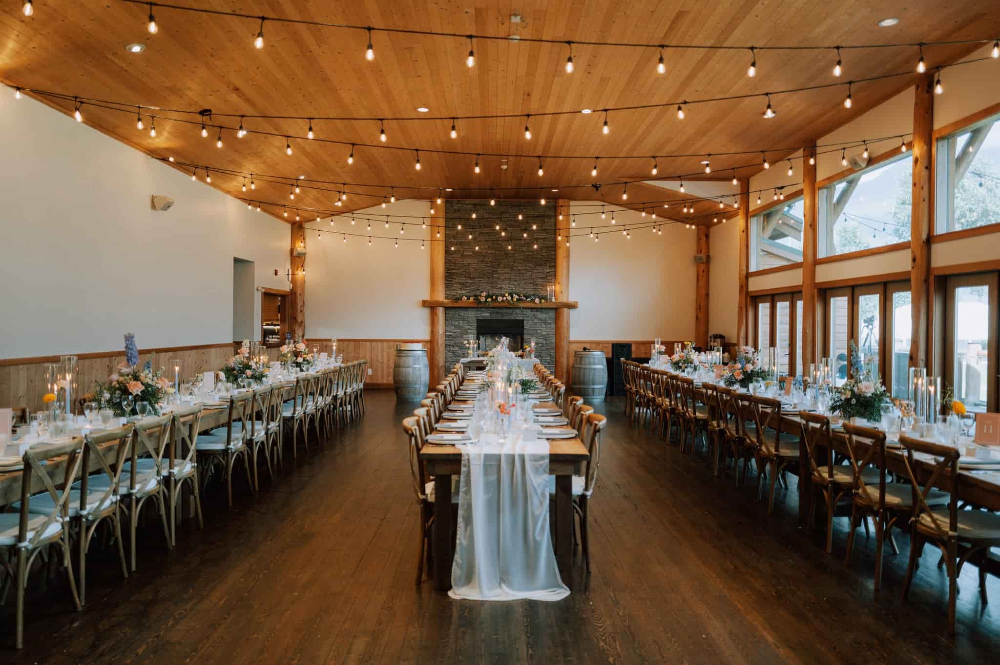 Rustic wedding reception in the Fraser River Lodge hall with wooden farmhouse tables, cross-back chairs, pastel florals and string lights